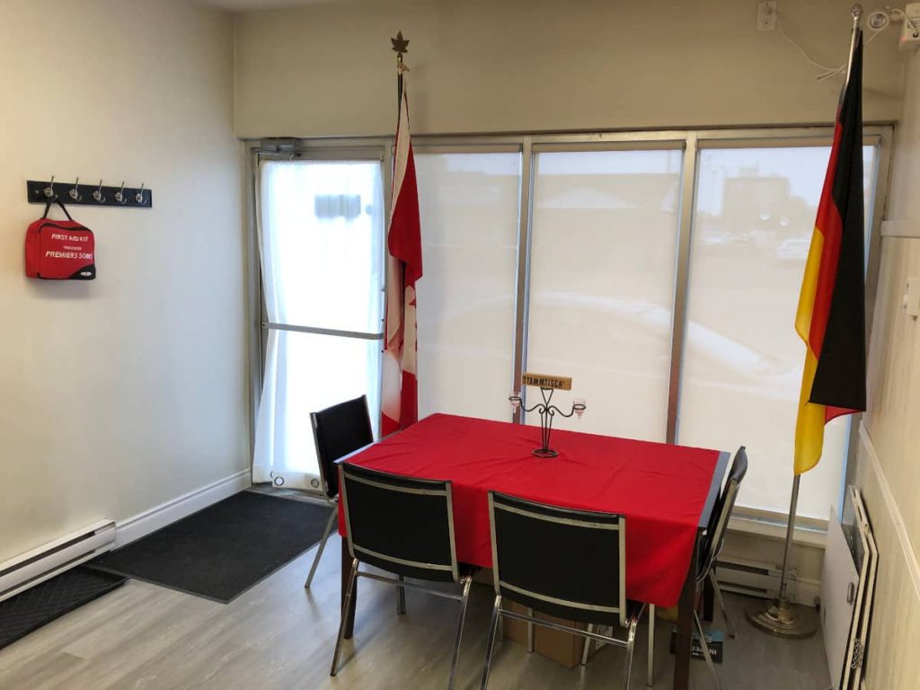 Interior view of our new premises on Franklin, showing another table with red tablecloth and 4 chairs, our Canadian and German flags and front door and big window with semi-transparent privacy paint through which we can kind of see the outside